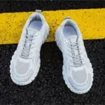 A pair of OEM Trendy Tire Sole Men Shoes—light, anti-slip, wear-resistant sneakers with cotton mesh and EVA—is placed side by side on black asphalt with a thick yellow line running underneath.