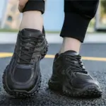 A person wearing black OEM Trendy Tire Sole Men Shoes—light, anti-slip, wear-resistant sneakers with rugged soles—is standing on an asphalt road.