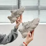 Two hands hold up Women's Low Top Mesh Sneakers with thick, height-increasing soles in front of a white wall and a window with closed blinds.