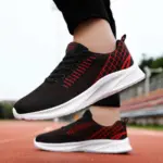 A person wearing Men’s Casual Sneakers—black mesh, lightweight, flying weaving lace-up with white EVA soles—stands on a running track, ideal for OEM/ODM sportswear brands prioritizing quality and style.