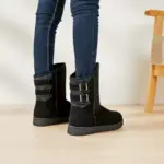 A person in dark skinny jeans and black Wholesale Fashion Western Designer Snow Boots for Women 2024 stands on a light wooden floor next to a wooden chair.