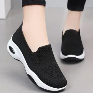 A person wears Fashion Slip on Custom Mesh Sneakers Sports Women Running Shoes 2024—black, with white anti-slip soles, knitted mesh texture, and a thick sole featuring a visible air cushion for extra comfort—standing on a light gray floor.