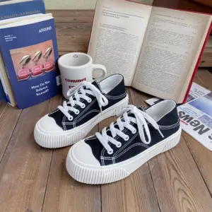 A pair of Breathable Designed Women Shoes Solid Cotton Sneakers—slip-on, low top canvas shoes—rests on a wooden surface, surrounded by books, a coffee mug, and newspapers.
