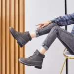A person sits on a chair with legs crossed, wearing rolled-up jeans, a plaid shirt, and Women Snow High Top Mountain Climbing Ankle Boots—outdoor leather anti-slip ankle boots for women—against a light background with wooden paneling.