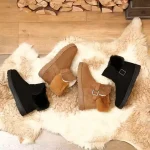 Two pairs of Warm Comfort Suede Boots—one black, one brown with fur trim and buckles—rest on a cream sheepskin rug on a wooden floor, firewood behind. Ideal for indoor/outdoor use; OEM/ODM customization available. Wholesale faux fur winter fashion boots.