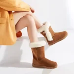 A person sits on a white surface wearing Hot Selling Winter Leather Boots Ankle Shoes—fashionable brown suede boots with white fur lining and decorative buttons, perfect for women seeking stylish winter footwear.