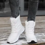 A person in Wholesale Wear Resistant Outdoor Cotton Snow Boots PU High Top Round Toe Rubber Women's Shoes and dark jeans stands on a wooden surface lightly dusted with snow.