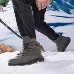 A person in High Quality 2024 Men Custom Lace up Ankle Snow Boots and black pants, using an OEM ODM trekking pole, walks on snow with snowy mountains in the background.